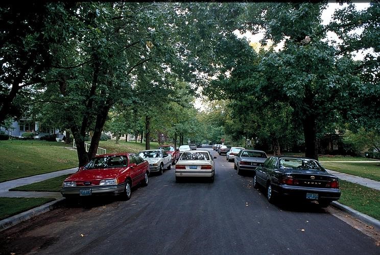 A narrow tree-lined street with many parked cars.